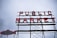 A large sign with the words 'PUBLIC MARKET' in bold red neon letters, supported by a metal framework. To the left, there is an additional sign shaped like a fish with the text 'CITY FISH MARKET'. The background is a cloudy sky, adding a moody atmosphere to the scene.