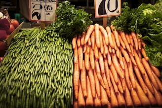 A display of green beans and carrots is positioned side by side on a market stall. The green beans are stacked neatly on the left, while the carrots are organized in a similar fashion on the right. Price signs are visible above the vegetables.
