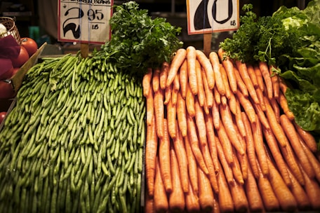 A display of green beans and carrots is positioned side by side on a market stall. The green beans are stacked neatly on the left, while the carrots are organized in a similar fashion on the right. Price signs are visible above the vegetables.