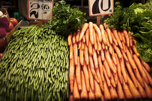 A display of green beans and carrots is positioned side by side on a market stall. The green beans are stacked neatly on the left, while the carrots are organized in a similar fashion on the right. Price signs are visible above the vegetables.