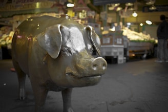 A large bronze pig statue is prominently displayed in an indoor market setting, surrounded by produce and market stalls. The atmosphere is bustling, with blurred figures of people engaged in shopping activities.