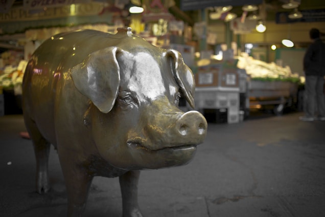 A large bronze pig statue is prominently displayed in an indoor market setting, surrounded by produce and market stalls. The atmosphere is bustling, with blurred figures of people engaged in shopping activities.