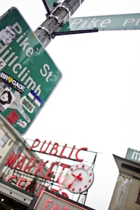 An angled view of a street sign adorned with various stickers, including one with the text 'Pike St Hillclimb', and another street sign labeled 'Pike Place'. In the background, the iconic red neon sign of the 'Public Market Center' is visible, along with its clock. The scene captures an urban area with overlapping signage and a slightly cloudy sky.