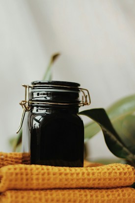 A dark glass jar with a metal clasp sits on a folded mustard-yellow waffle-textured cloth. In the background, there are blurred green leaves against a neutral wall, giving a natural and cozy atmosphere.