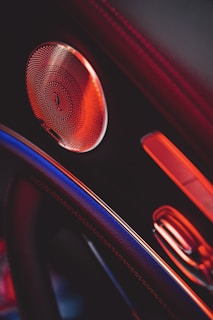 A close-up view of a car interior featuring a speaker with intricate detailing, highlighted by ambient red lighting. Visible elements include part of the steering wheel and a section of the door panel.