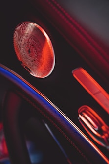 A close-up view of a car interior featuring a speaker with intricate detailing, highlighted by ambient red lighting. Visible elements include part of the steering wheel and a section of the door panel.