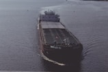 A large cargo ship is sailing on open water. The ship has a dark hull with visible markings and appears to be carrying a large number of shipping containers on its deck. The water around the ship is calm, reflecting the light of the overcast sky.