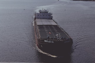 A large cargo ship is sailing on open water. The ship has a dark hull with visible markings and appears to be carrying a large number of shipping containers on its deck. The water around the ship is calm, reflecting the light of the overcast sky.