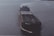 A large cargo ship is sailing on open water. The ship has a dark hull with visible markings and appears to be carrying a large number of shipping containers on its deck. The water around the ship is calm, reflecting the light of the overcast sky.