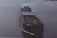 A large cargo ship is sailing on open water. The ship has a dark hull with visible markings and appears to be carrying a large number of shipping containers on its deck. The water around the ship is calm, reflecting the light of the overcast sky.