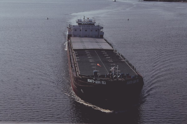 A large cargo ship is sailing on open water. The ship has a dark hull with visible markings and appears to be carrying a large number of shipping containers on its deck. The water around the ship is calm, reflecting the light of the overcast sky.