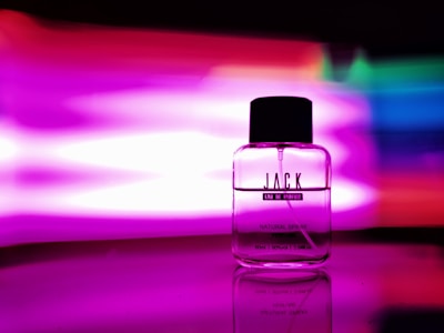 A glass perfume bottle labeled 'JACK Eau de Parfum' with a black cap is placed against a vibrant multicolored background with prominent pink and purple hues. The reflective surface underneath enhances the vivid colors and sleek design of the bottle.