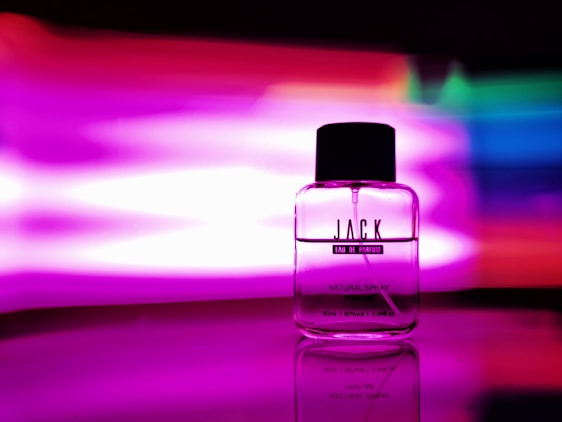 A glass perfume bottle labeled 'JACK Eau de Parfum' with a black cap is placed against a vibrant multicolored background with prominent pink and purple hues. The reflective surface underneath enhances the vivid colors and sleek design of the bottle.