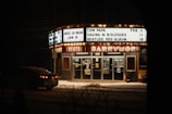 A nighttime scene featuring a theater with a brightly lit marquee displaying upcoming events and performance dates. Posters are visible on the theater doors and a car is parked on the snow-covered street in front of it. The atmosphere is quiet with a hint of nostalgia.