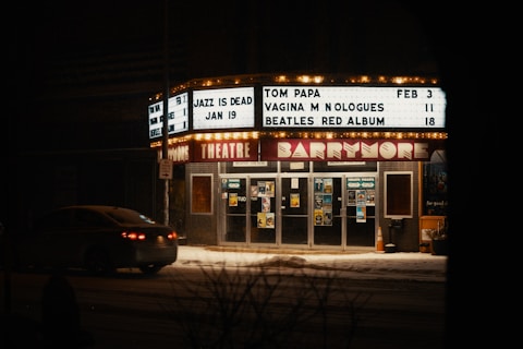 A nighttime scene featuring a theater with a brightly lit marquee displaying upcoming events and performance dates. Posters are visible on the theater doors and a car is parked on the snow-covered street in front of it. The atmosphere is quiet with a hint of nostalgia.