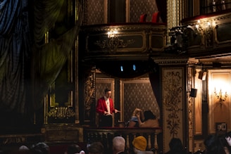 An ornate theater interior with lavish decorations in golden hues surrounds a balcony adorned with intricate patterns. A person dressed in a bright red jacket stands on the balcony, engaging with two seated individuals. The opulent setting features chandeliers and elaborate molding, creating a rich and classic atmosphere.
