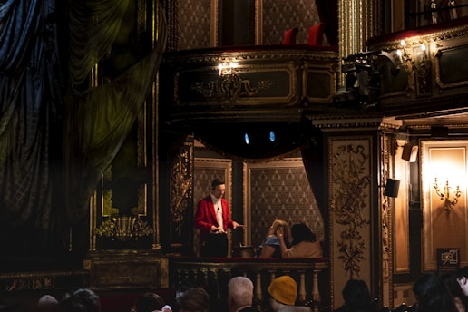 An ornate theater interior with lavish decorations in golden hues surrounds a balcony adorned with intricate patterns. A person dressed in a bright red jacket stands on the balcony, engaging with two seated individuals. The opulent setting features chandeliers and elaborate molding, creating a rich and classic atmosphere.