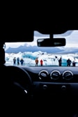 A scenic view of icebergs and a glacier seen through a car's windshield. A group of people stand on the icy landscape, dressed in warm clothing. The dashboard and steering wheel of the car are visible in the foreground.