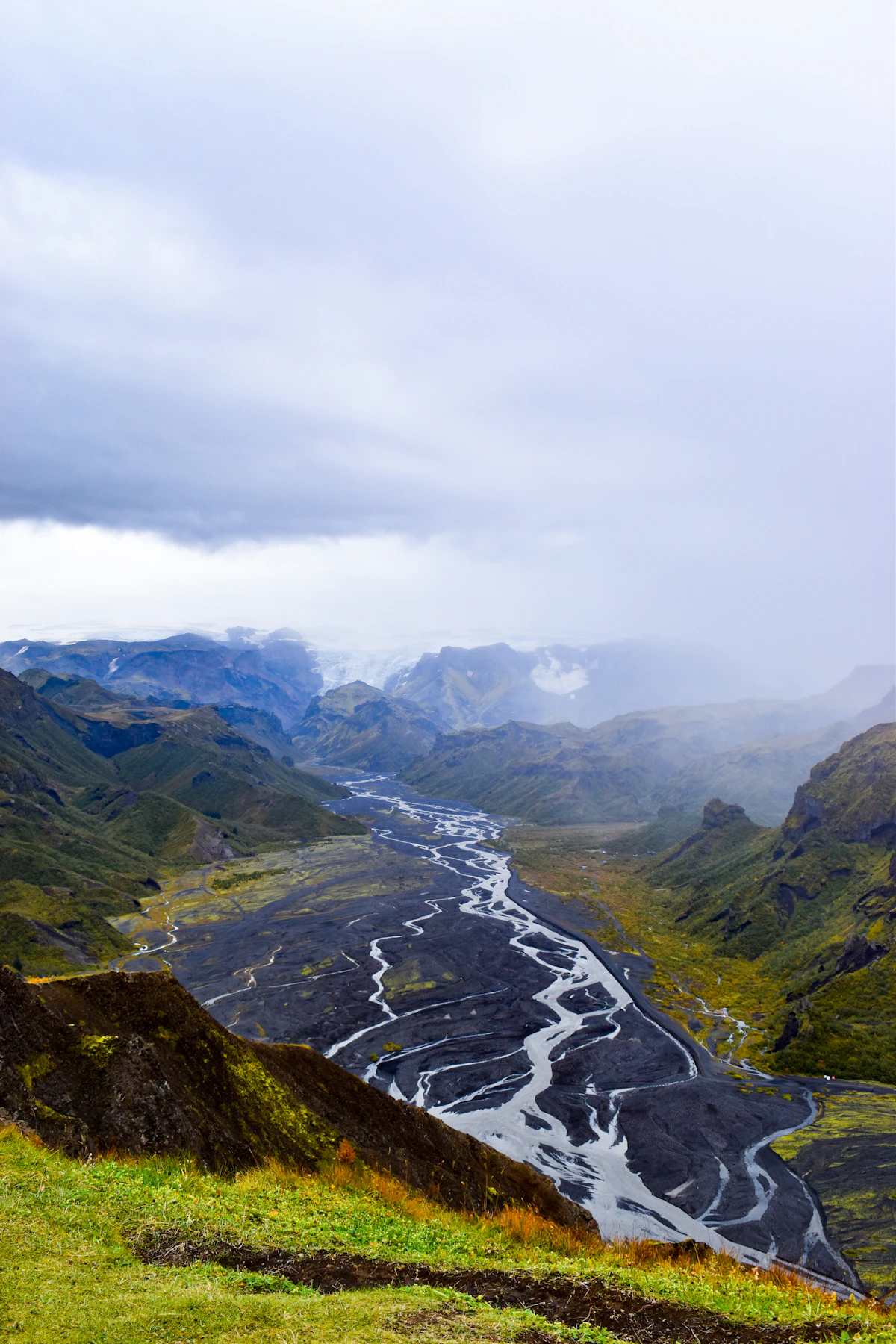 A braided glacial river in a wide gravel valley between green ridges at Þórsmörk, under low cloud