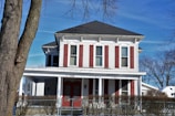 A two-story house with white siding and red shutters, surrounded by leafless trees in a winter setting. The house features a large porch with columns and a chain-link fence in front. Snow is visible on the ground, and the sky is clear and blue.