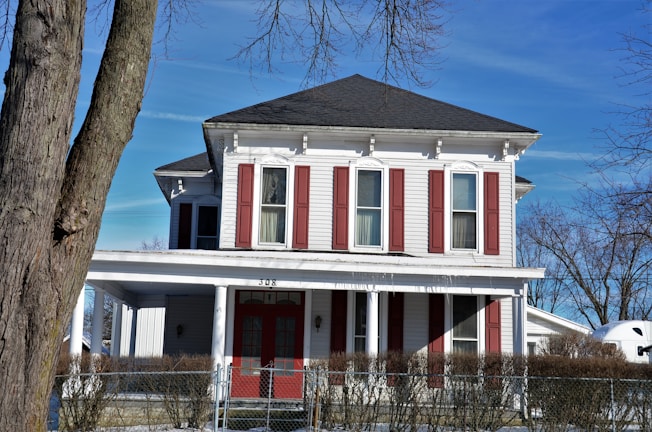 A two-story house with white siding and red shutters, surrounded by leafless trees in a winter setting. The house features a large porch with columns and a chain-link fence in front. Snow is visible on the ground, and the sky is clear and blue.