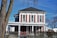A two-story house with white siding and red shutters, surrounded by leafless trees in a winter setting. The house features a large porch with columns and a chain-link fence in front. Snow is visible on the ground, and the sky is clear and blue.