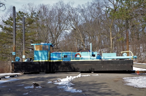 A large blue and black industrial machine with various components, positioned on a paved road in a wooded area during winter. The ground is partially covered with patches of snow. Tall trees without leaves are in the background, indicating a cold season.