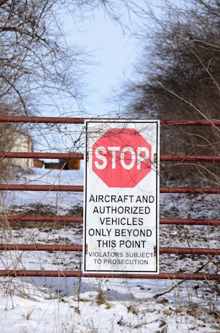 A warning sign is attached to a metal gate surrounded by trees and snow. The sign has a red stop symbol and text indicating restricted access for aircraft and authorized vehicles only, with a warning about prosecution for violators. The ground is covered with a light layer of snow, and bare branches are visible around.