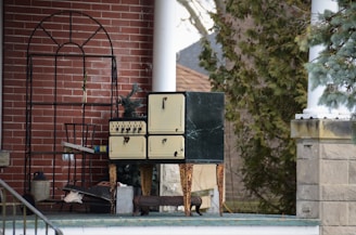 A vintage oven with cream-colored doors and black detailing stands on a porch next to a black metal structure. The background consists of a brick wall and some evergreen trees. Various objects, including a metal jug and pieces of wood, are scattered around.