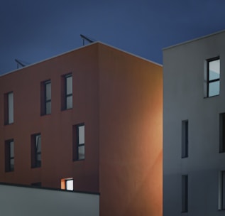 Two modern apartment buildings are shown against a dark, evening sky. One building is a muted orange with rectangular windows, some of which are illuminated from within. The other building is a light grey with similar window designs, contributing to the minimalist, geometric aesthetic.