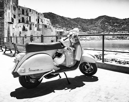 A vintage scooter is parked on a terrace overlooking a coastal town. In the background, there are buildings along with hilly terrain, and the sea is visible with sun umbrellas lined up on the beach. The image is in black and white, giving it a classic, timeless feel.
