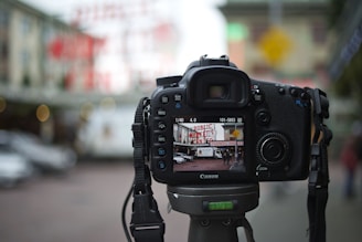 A DSLR camera is mounted on a tripod in an outdoor setting, focusing on a scene displaying a market sign and people in the background. The LCD screen shows an in-focus image of a street view with a market, cars, and pedestrians, while the background is blurred.