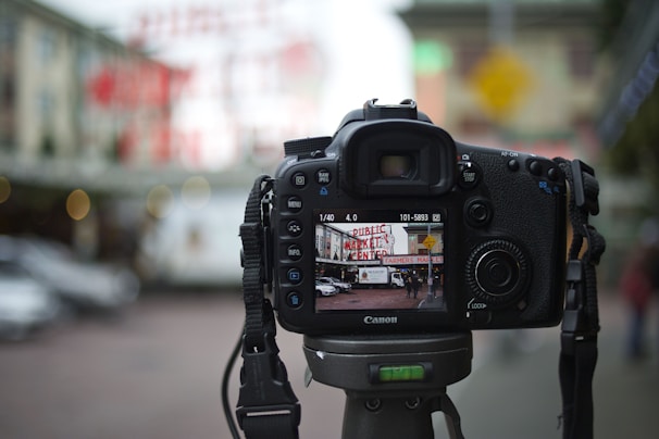 A DSLR camera is mounted on a tripod in an outdoor setting, focusing on a scene displaying a market sign and people in the background. The LCD screen shows an in-focus image of a street view with a market, cars, and pedestrians, while the background is blurred.