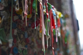 A wall covered with numerous pieces of colorful chewing gum in various shapes and sizes, some hanging and others pressed flat against the surface. The image captures a close-up view showing the texture and vivid colors against the background.