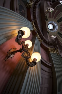 An ornate interior featuring an intricately designed hanging lamp with three glowing orbs, set against large, fluted columns and decorative molding. The scene includes a circular skylight above, casting natural light into the space, accentuating the architectural elegance and historical ambiance.