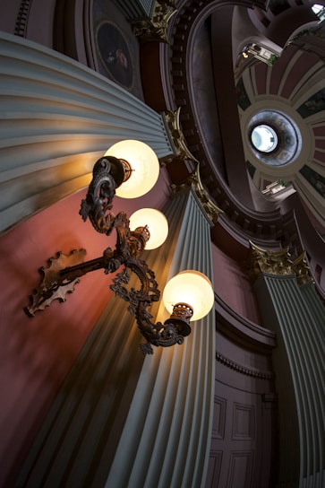 An ornate interior featuring an intricately designed hanging lamp with three glowing orbs, set against large, fluted columns and decorative molding. The scene includes a circular skylight above, casting natural light into the space, accentuating the architectural elegance and historical ambiance.