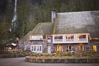 A large stone building with a steep gabled roof, adorned with string lights creating a warm and inviting atmosphere. The structure features multiple windows and a chimney, surrounded by lush forest with a waterfall visible in the background.