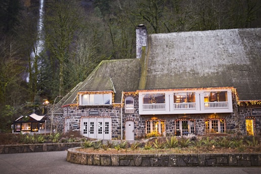 A large stone building with a steep gabled roof, adorned with string lights creating a warm and inviting atmosphere. The structure features multiple windows and a chimney, surrounded by lush forest with a waterfall visible in the background.