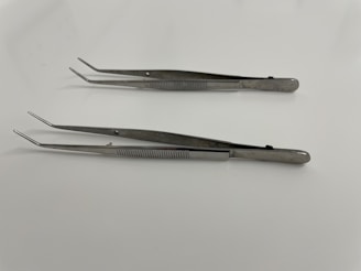 Two stainless steel tweezers are laid out on a smooth, white surface. Both tweezers have a straight, elongated design with slightly pointed tips and textured grips along the handles.