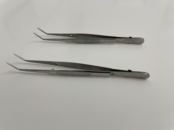 Two stainless steel tweezers are laid out on a smooth, white surface. Both tweezers have a straight, elongated design with slightly pointed tips and textured grips along the handles.