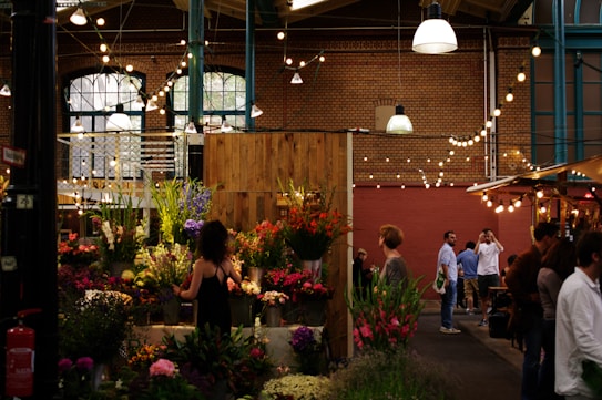 A bustling indoor market with an array of colorful flowers and plants. Several people stroll past, some interacting with the vendors, others observing the displays. The high ceiling is adorned with strings of round lights, and the brick walls and structured beams add a rustic charm to the setting.