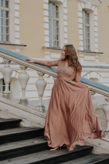 A stylish model wearing a flowing rose gold silk dress standing on a sunlit terrace.