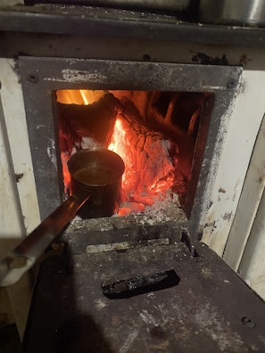 A metal container with a long handle rests inside a wood-burning stove. The stove is filled with glowing embers and red-hot logs that emit a bright, warm glow. The area surrounding the open stove is dark and slightly worn from use, highlighting the intensity and focus of the internal fire.