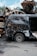 A damaged black van with multiple bullet holes is parked in a scrapyard. Surrounding it are piles of rusted, crushed vehicles stacked high, creating a chaotic and cluttered scene. The ground is littered with debris, and the sky above is partly cloudy.