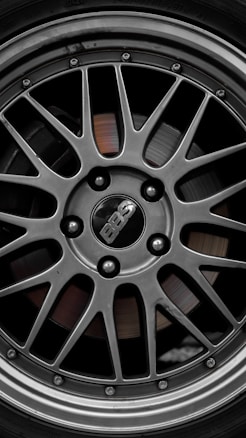 A close-up view of a metallic alloy wheel with a sleek, intricate multi-spoke design. The center cap displays the 'BBS' logo, surrounded by lug nuts securing the wheel in place. The polished surface gleams with a combination of silver and dark gray tones, accentuating the craftsmanship.