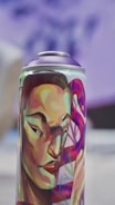 A spray can with vibrant and colorful artwork featuring a stylized human face with strong features. The face is adorned with bold colors including greens, reds, and purples, and an abstract background design complements the portrait.