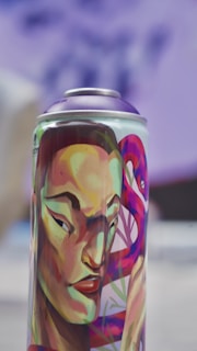 A spray can with vibrant and colorful artwork featuring a stylized human face with strong features. The face is adorned with bold colors including greens, reds, and purples, and an abstract background design complements the portrait.