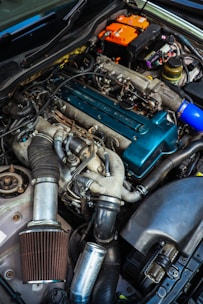A close-up view of a car engine with various parts visible, including a large air filter, hoses, and a blue valve cover. The image also shows an orange battery and numerous wires and components arranged around the engine bay.