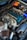 A close-up view of a car engine with various parts visible, including a large air filter, hoses, and a blue valve cover. The image also shows an orange battery and numerous wires and components arranged around the engine bay.