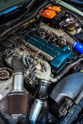 A close-up view of a car engine with various parts visible, including a large air filter, hoses, and a blue valve cover. The image also shows an orange battery and numerous wires and components arranged around the engine bay.